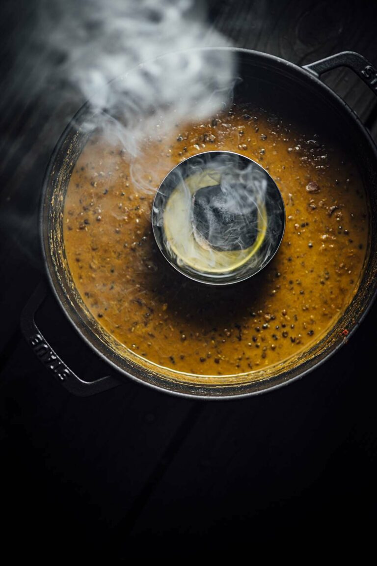 Overhead view of dal makhani in a black Dutch oven with a small metal bowl of smoking charcoal in the center, demonstrating the dhungar smoking technique.