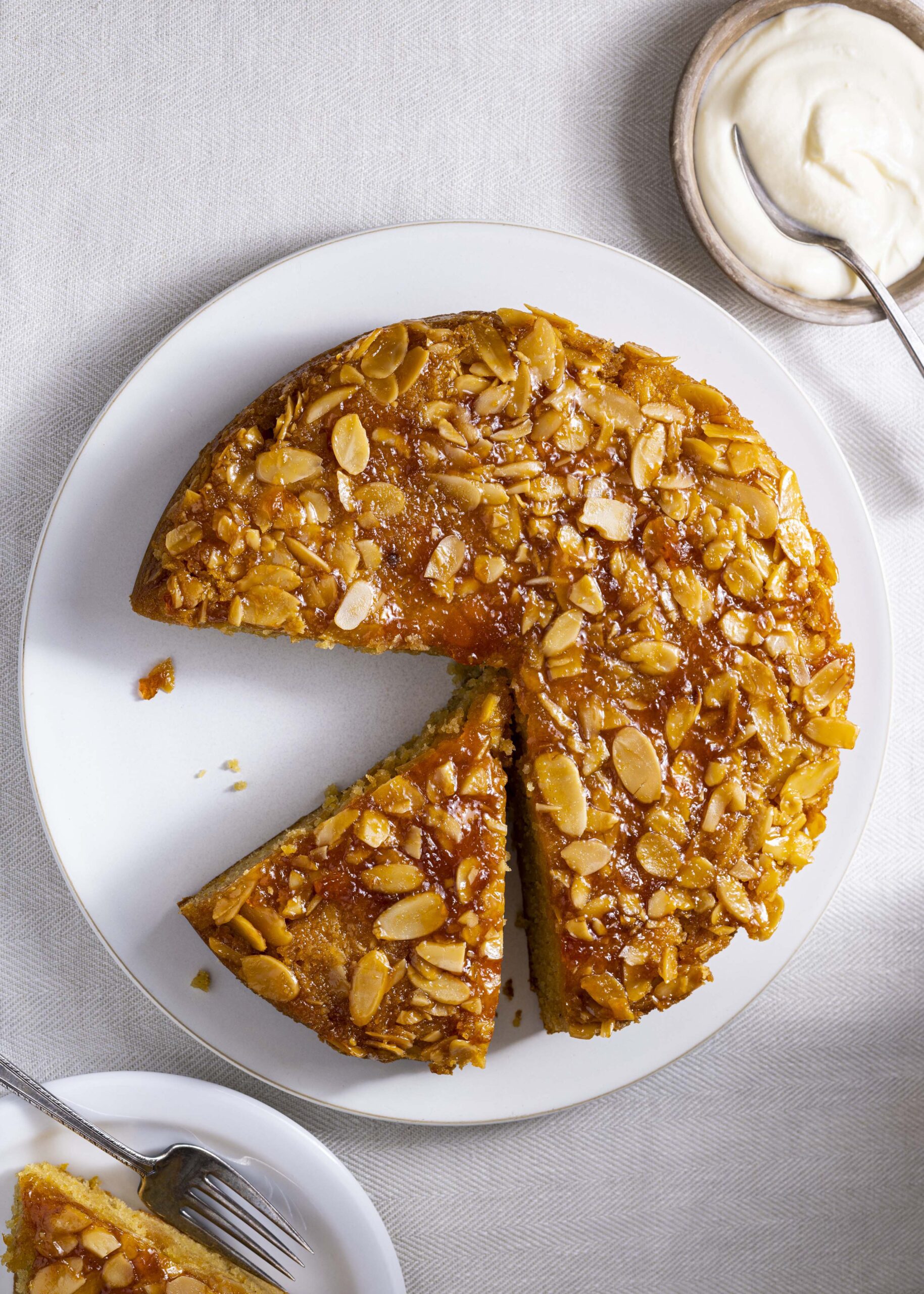 Almond apricot honey cake served on a white plate with a slice cut, showing the caramelized apricot and toasted almond topping, with crème fraîche on the side