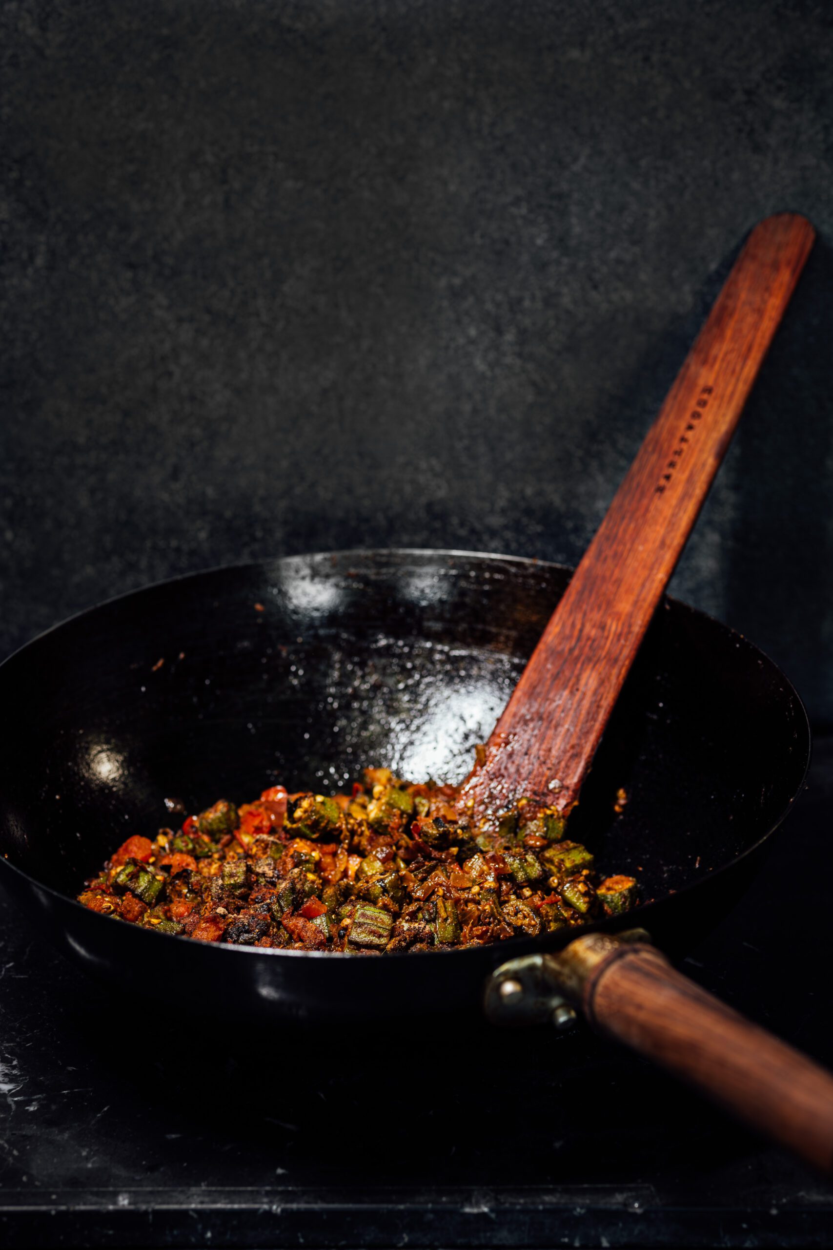Bhindi masala with caramelized okra, tomato, and spices in a black carbon steel wok with a wooden spatula.