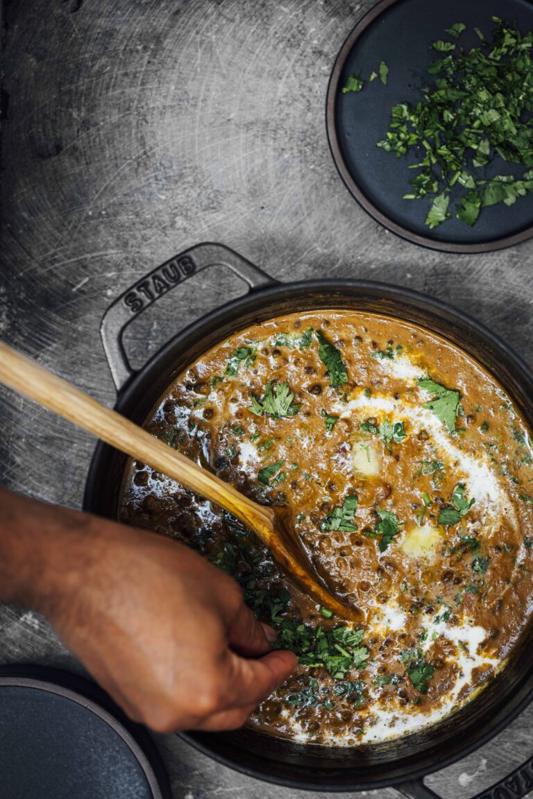 Overhead view of dal makhani in a black Staub Dutch oven, garnished with cream, a pat of butter, and fresh cilantro, stirred with a wooden spoon.