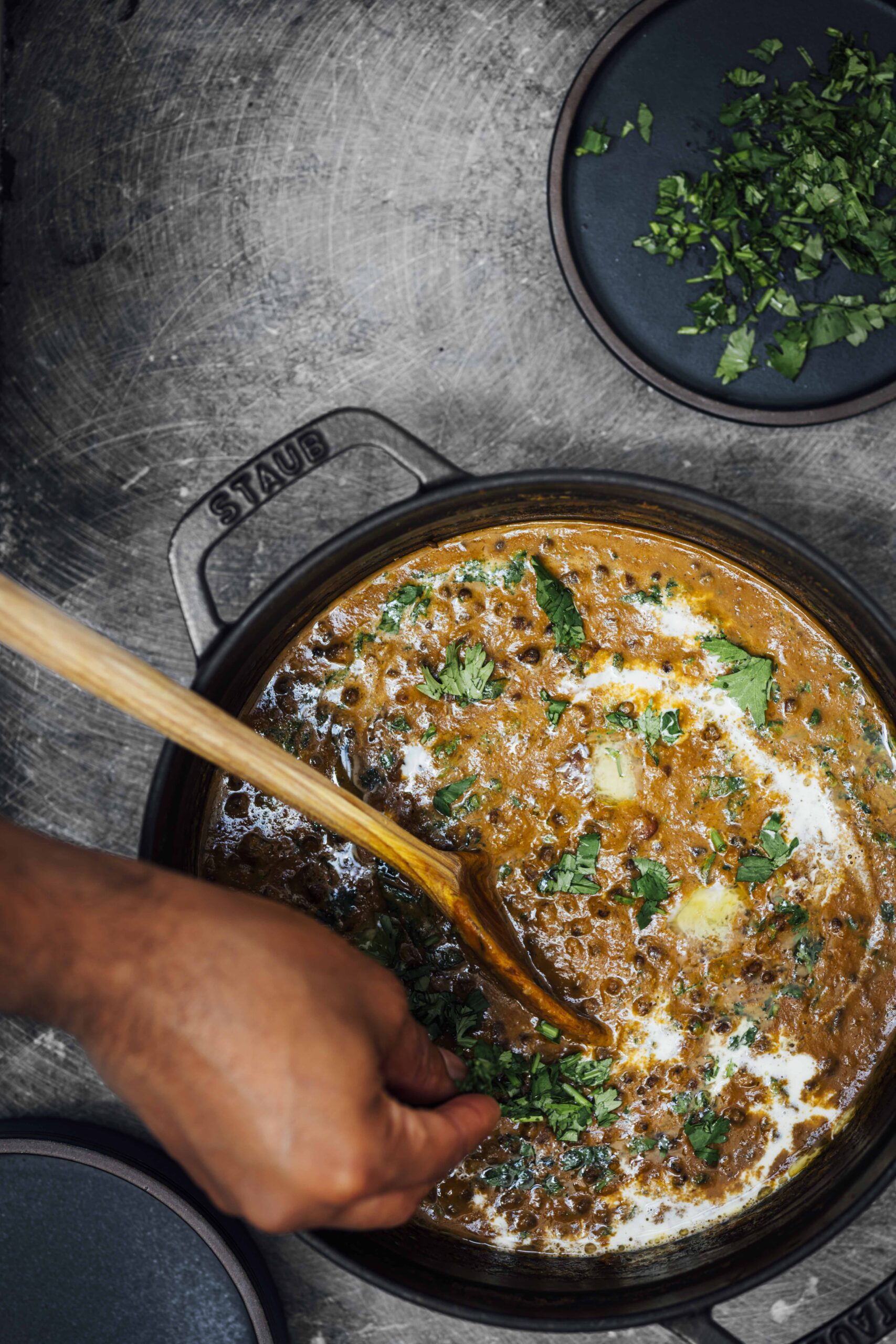 Overhead view of dal makhani in a black Staub Dutch oven, garnished with cream, a pat of butter, and fresh cilantro, stirred with a wooden spoon.