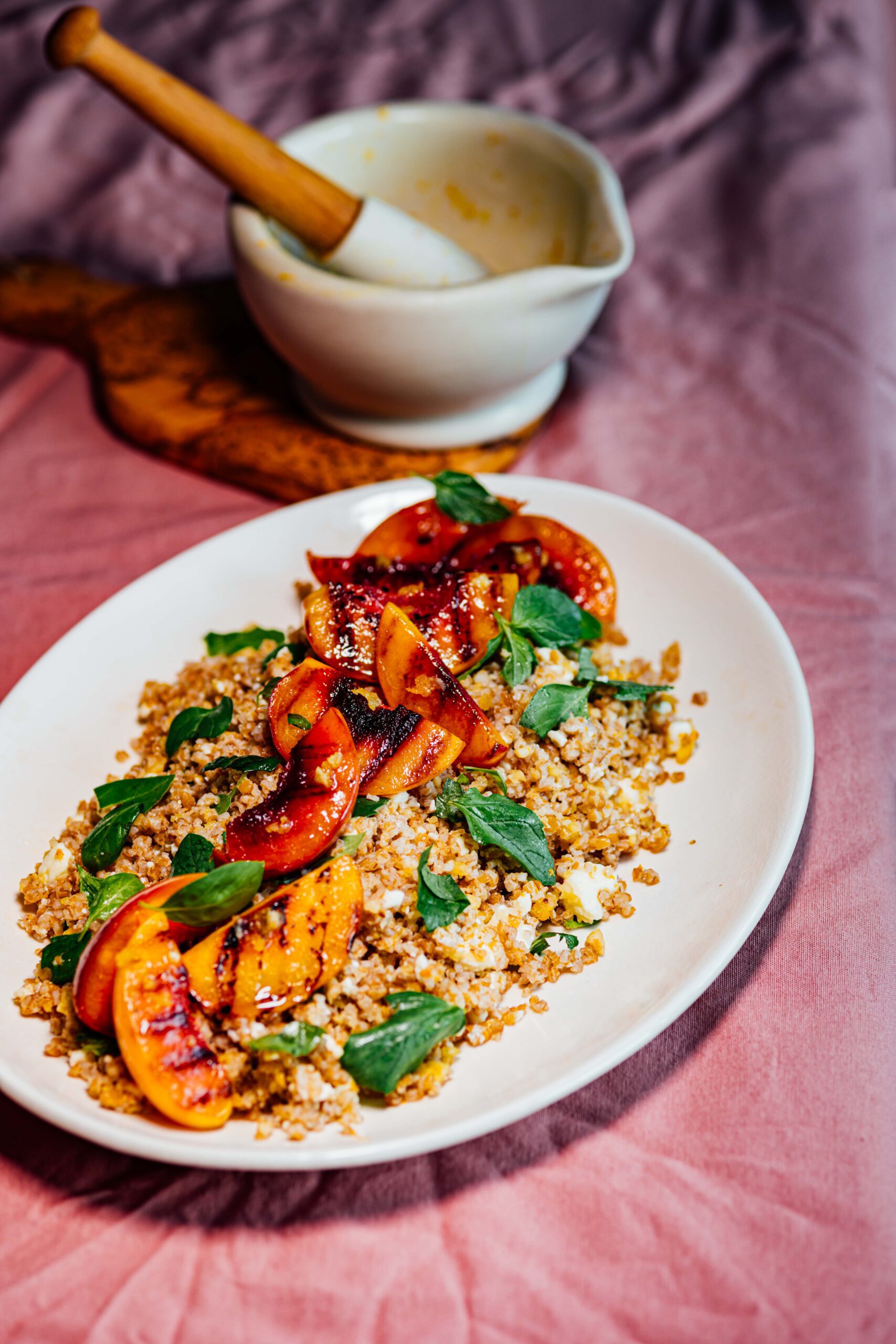 White oval plate of bulgur salad topped with charred grilled nectarine wedges and fresh mint leaves, with a mortar and pestle in the background
